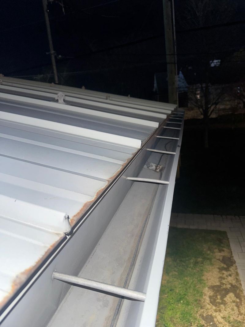 Gros plan pris de nuit, au flash&nbsp;: l’intérieur d’une gouttière d’aluminium, complètement vide, le long du bord d’un toit de tôle.