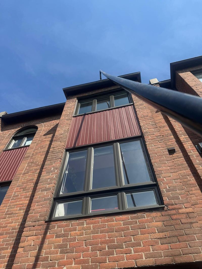 La perche noire monte en diagonale jusqu’à la gouttière noire d’une maison de ville en brique rouge. Ciel bleu foncé, ombre de la perche sur la brique.